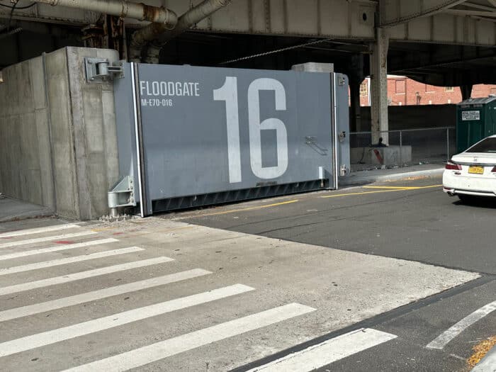 Large gray floodgate labeled "FLOODGATE MF-070-016 16" is installed under an overpass near a crosswalk and parked car.