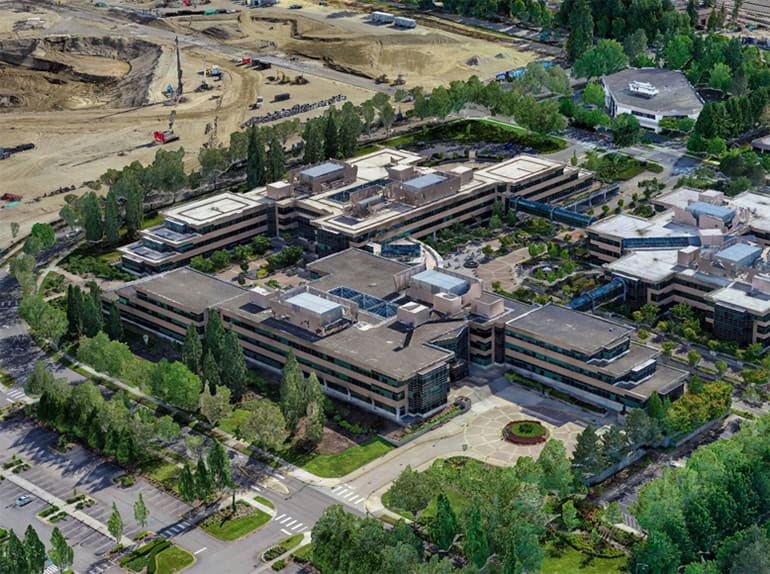Aerial view of a large office complex surrounded by trees, parking lots, and a nearby construction site using Intelligent Digital Twins for unified reality modeling and efficient earth-moving equipment deployment.