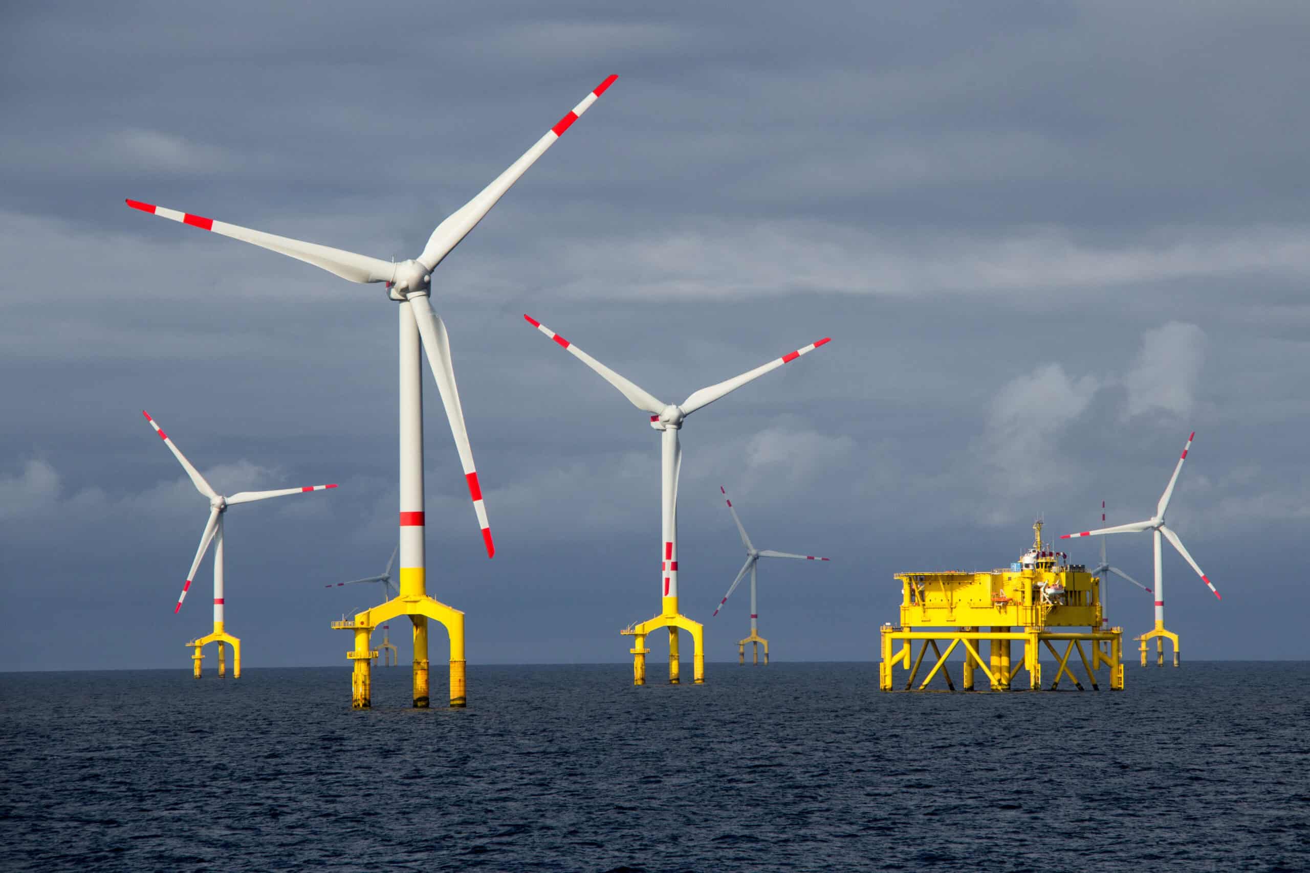 A wide view of an offshore wind farm with multiple wind turbines standing in the ocean, evenly spaced across the water. The turbines are tall with white blades and red safety stripes and bright yellow foundations, with rotating blades capturing wind energy. The sea is calm under a partially cloudy sky, and the horizon stretches into the distance.