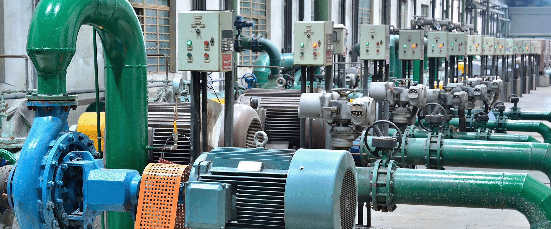 Row of industrial pumps and motors with green pipes and control panels in a factory setting, showcasing resilient water infrastructure designed for efficiency and reduced energy costs.