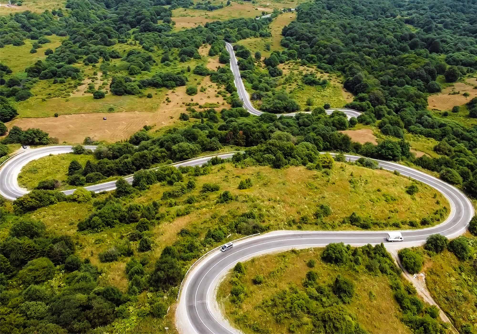 Aerial view of a curvy road winding through a green, hilly landscape with dense patches of trees and vegetation, showcasing intricate infrastructure design. Sparse traffic is visible on the road, maintained by state and local authorities.