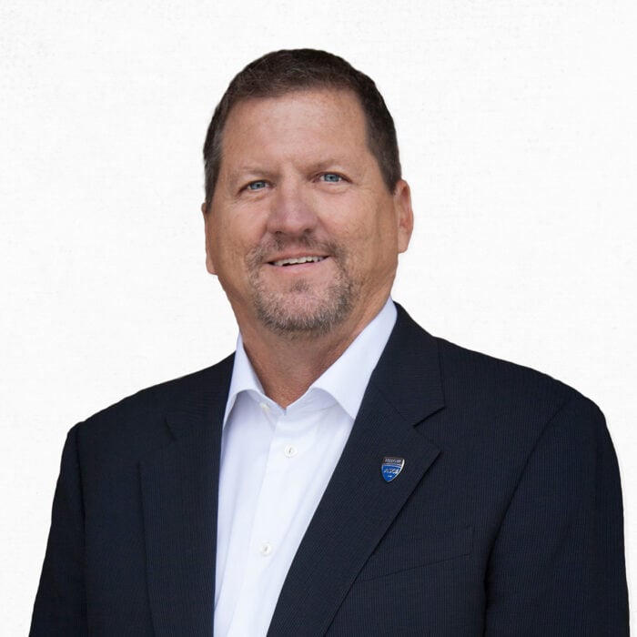 A man with short brown hair and a goatee wearing a dark suit jacket, white shirt, and a small pin, standing against a plain white background.