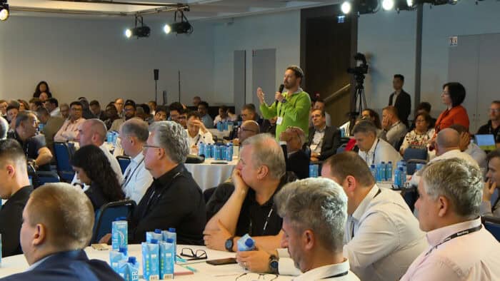 A group of people seated in a conference room, with one person standing and speaking, holding a microphone. Bottles of water and conference materials are on the tables.