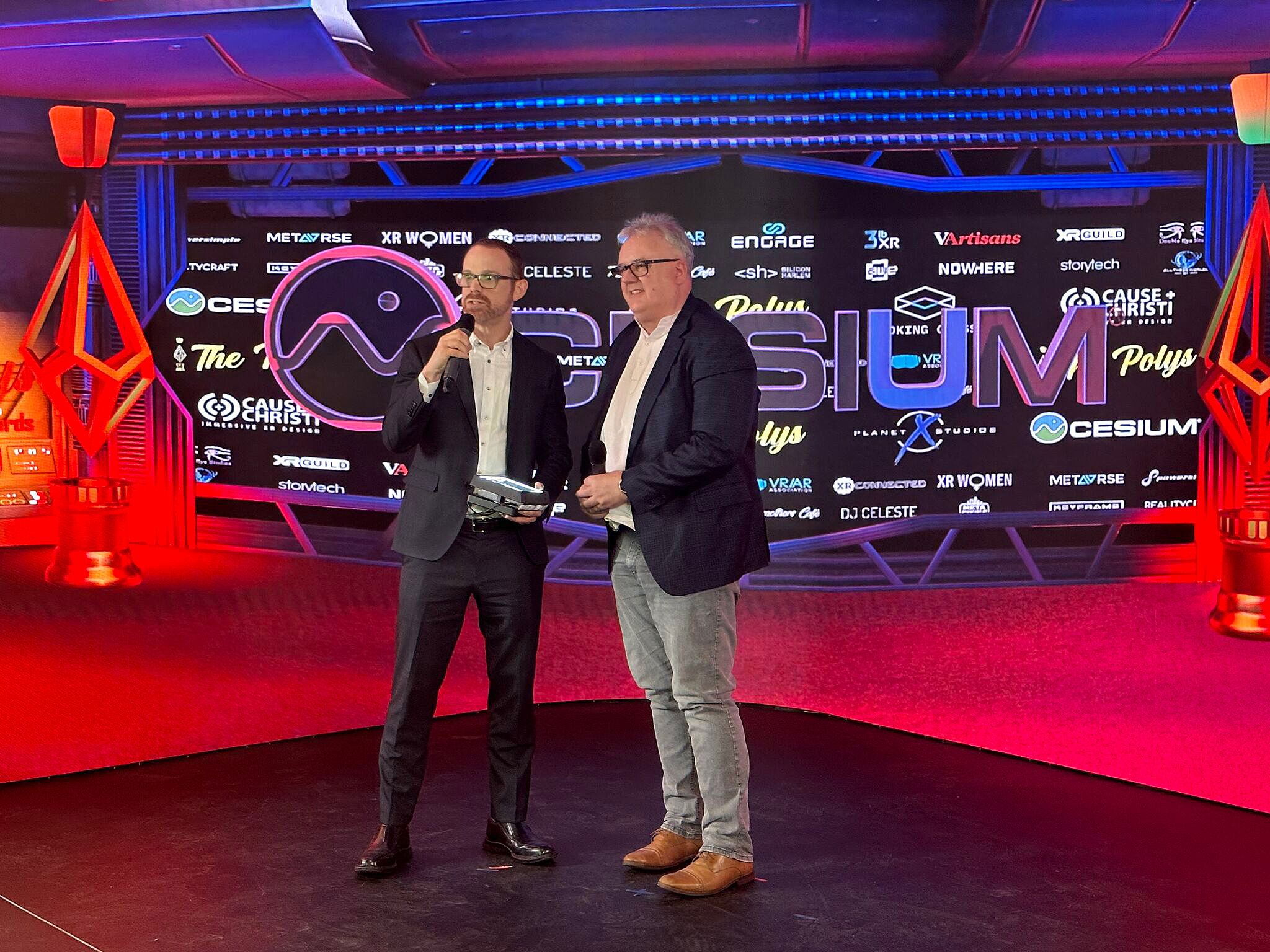 Patrick Cozzi and Neil Trevett on stage at The Polys Immersive Awards with a colorful digital background featuring the Cesium logo and other brands.