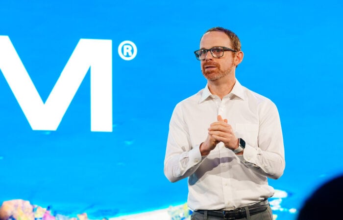 A person in a white shirt and glasses speaks on stage with a blue background and a partial letter "M" visible.