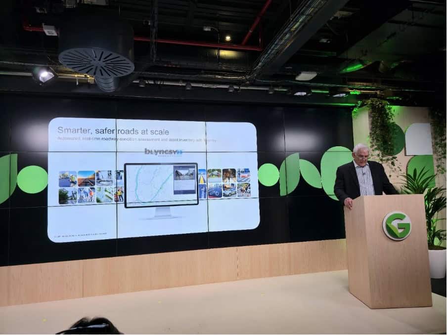 A man stands at a podium with a green "G" logo, presenting a slide titled "Smarter, safer roads at scale" in a modern conference room, highlighting how Google uses AI for Earth to predict disasters and enhance safety.