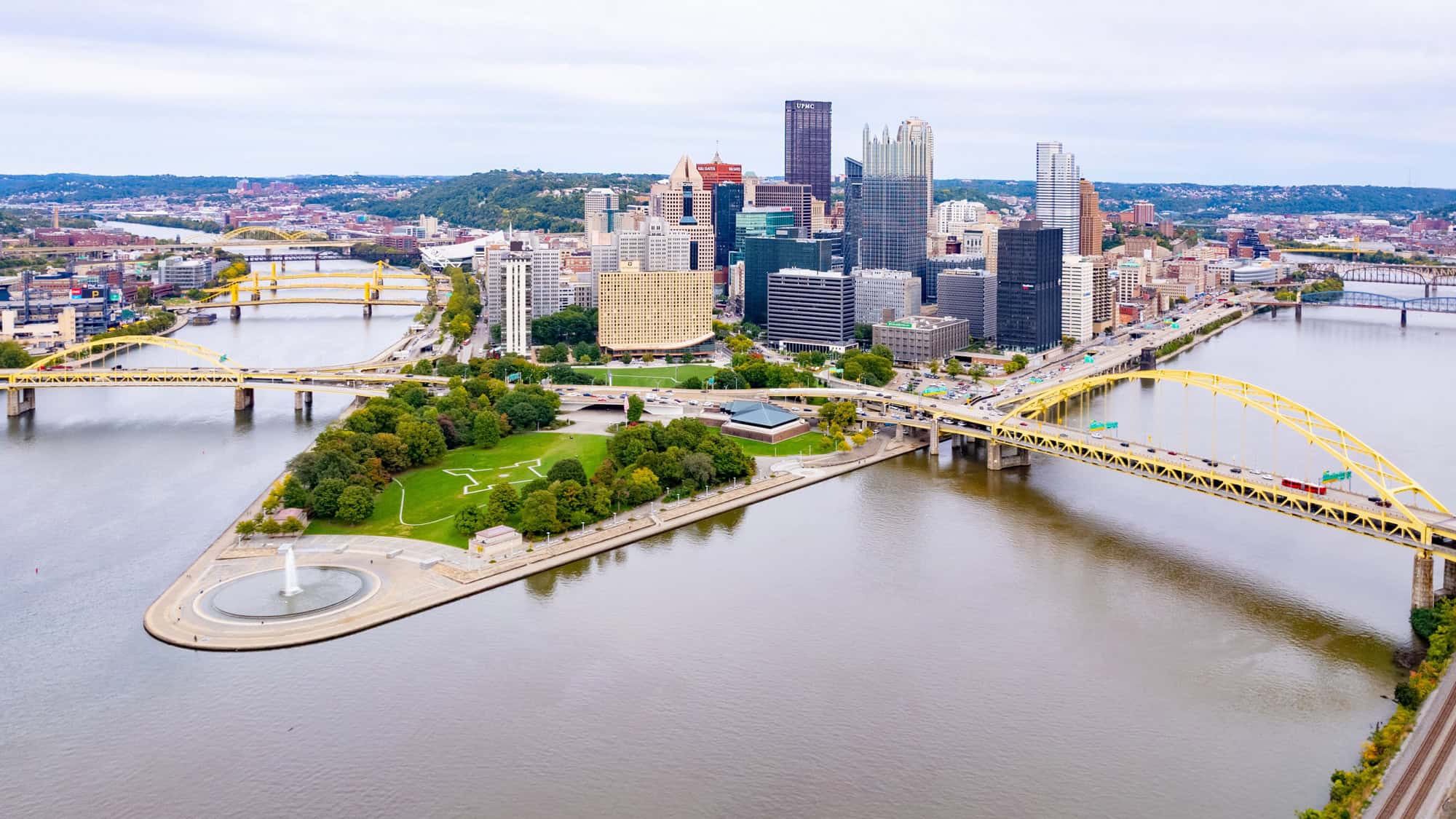 Aerial view of downtown Pittsburgh, featuring bridges, rivers, and Point State Park with its fountain at the confluence of the Allegheny, Monongahela, and Ohio Rivers.