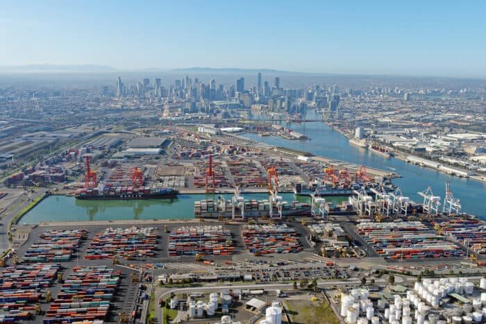 Aerial view of a large port with numerous shipping containers and cranes. A city skyline is visible in the background under a clear blue sky.