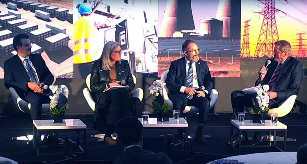 Four people in formal attire sit on a panel discussion stage with microphones, in front of an energy-themed backdrop featuring power plants and high-voltage lines.