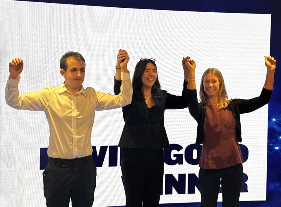 Three people stand on stage, holding hands up in celebration and smiling, after winning a global contest, with a large bright screen displaying partially visible text behind them.