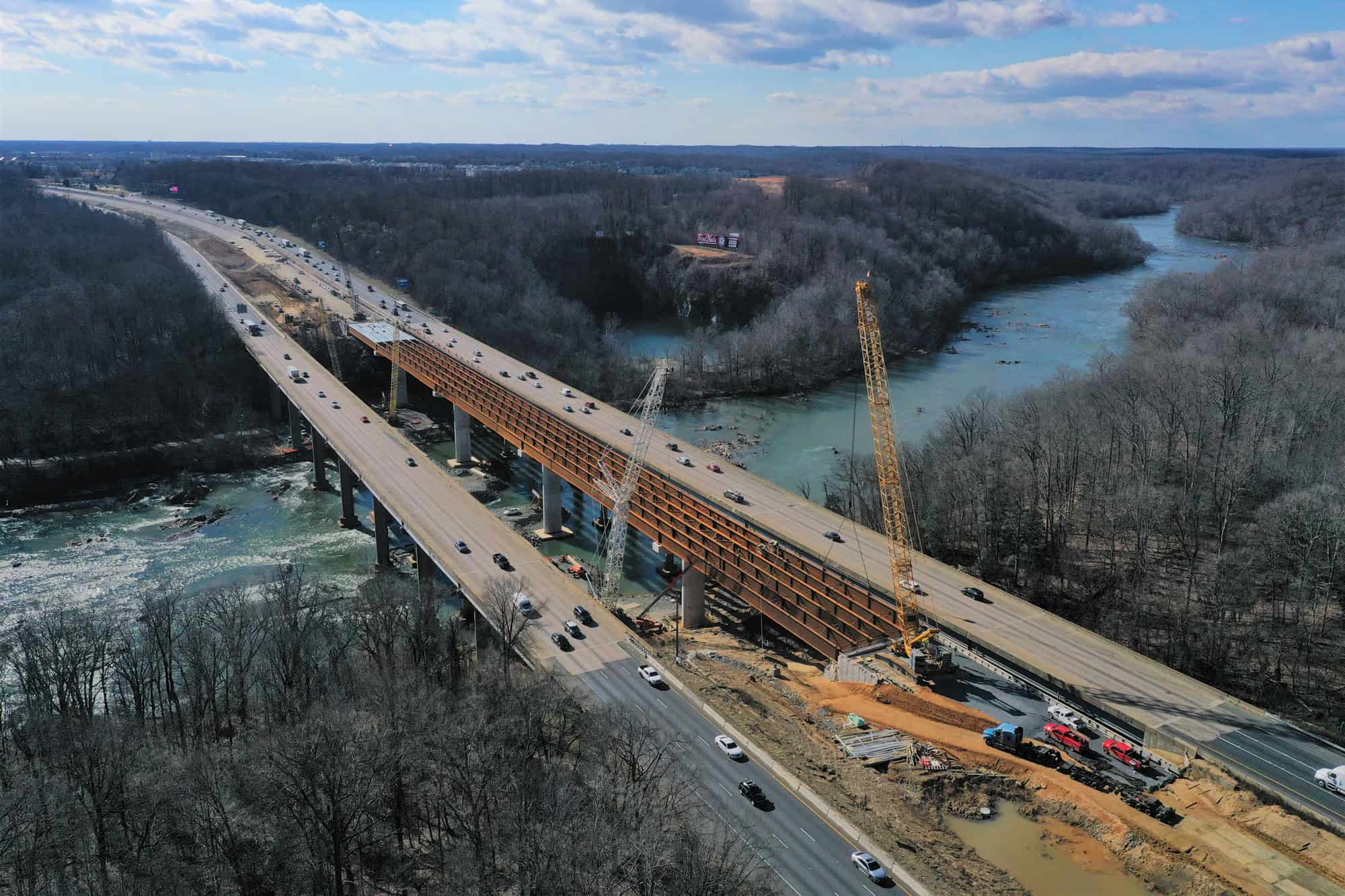 Aerial view of a bridge under construction over a river, with cranes, construction vehicles, and cars on the adjacent completed bridge.