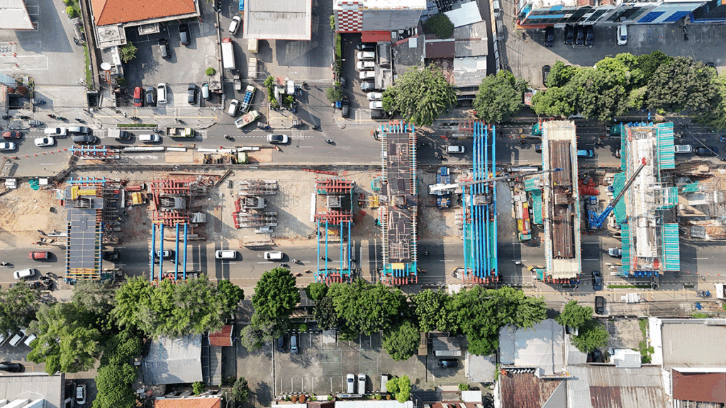 Aerial view of an urban construction site with several bridge sections being built over a busy road, surrounded by buildings and trees.