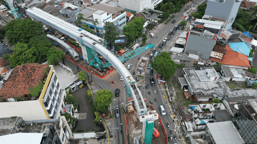 Aerial view of a city intersection with an elevated railway under construction, showing cranes, scaffolding, and vehicles moving on the surrounding roads.