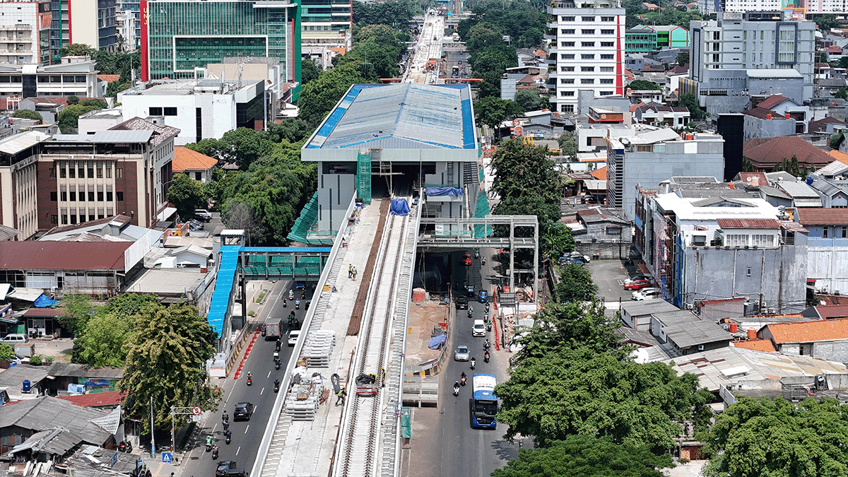 Aerial view of an elevated train station under construction in a dense urban area, with surrounding buildings and cars on the road below.