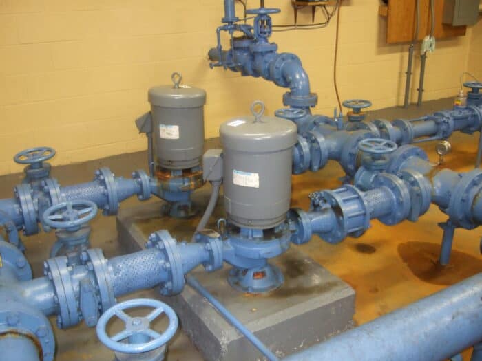 Blue industrial pipes and valves are seamlessly connected, incorporating variable frequency drives to enhance pump efficiency. Two large cylindrical units sit on a concrete base, highlighting the true costs and benefits of modern engineering solutions.