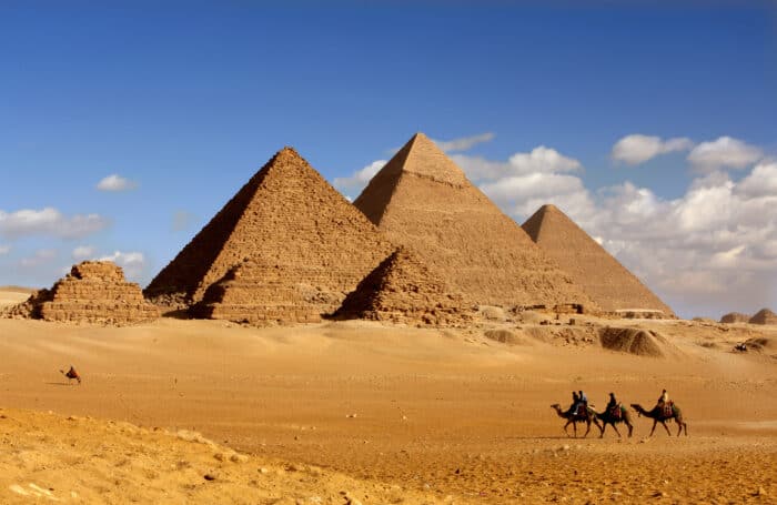 Three large pyramids and two smaller ones stand in a desert landscape with four people riding camels in the foreground under a blue sky with scattered clouds.