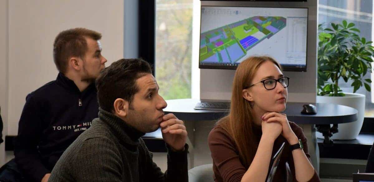 image of three individuals are attentively looking at a computer screen displaying a colorful architectural 3D model in an office setting with a large window, a potted plant, and advanced software contributing to their éxito.