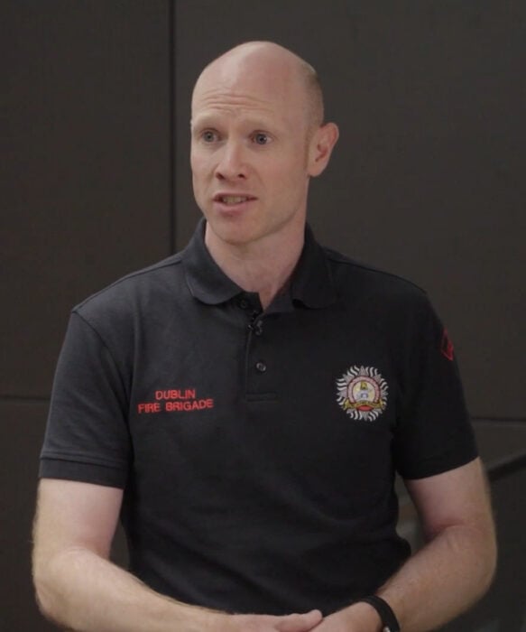 A man in a black "Dublin Fire Brigade" polo shirt stands indoors, speaking with a neutral expression.