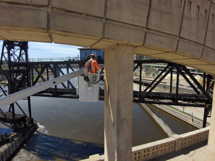 A worker in a bucket lift inspects or repairs the side of the large concrete Robert Street Bridge over a river, with another bridge visible in the background.