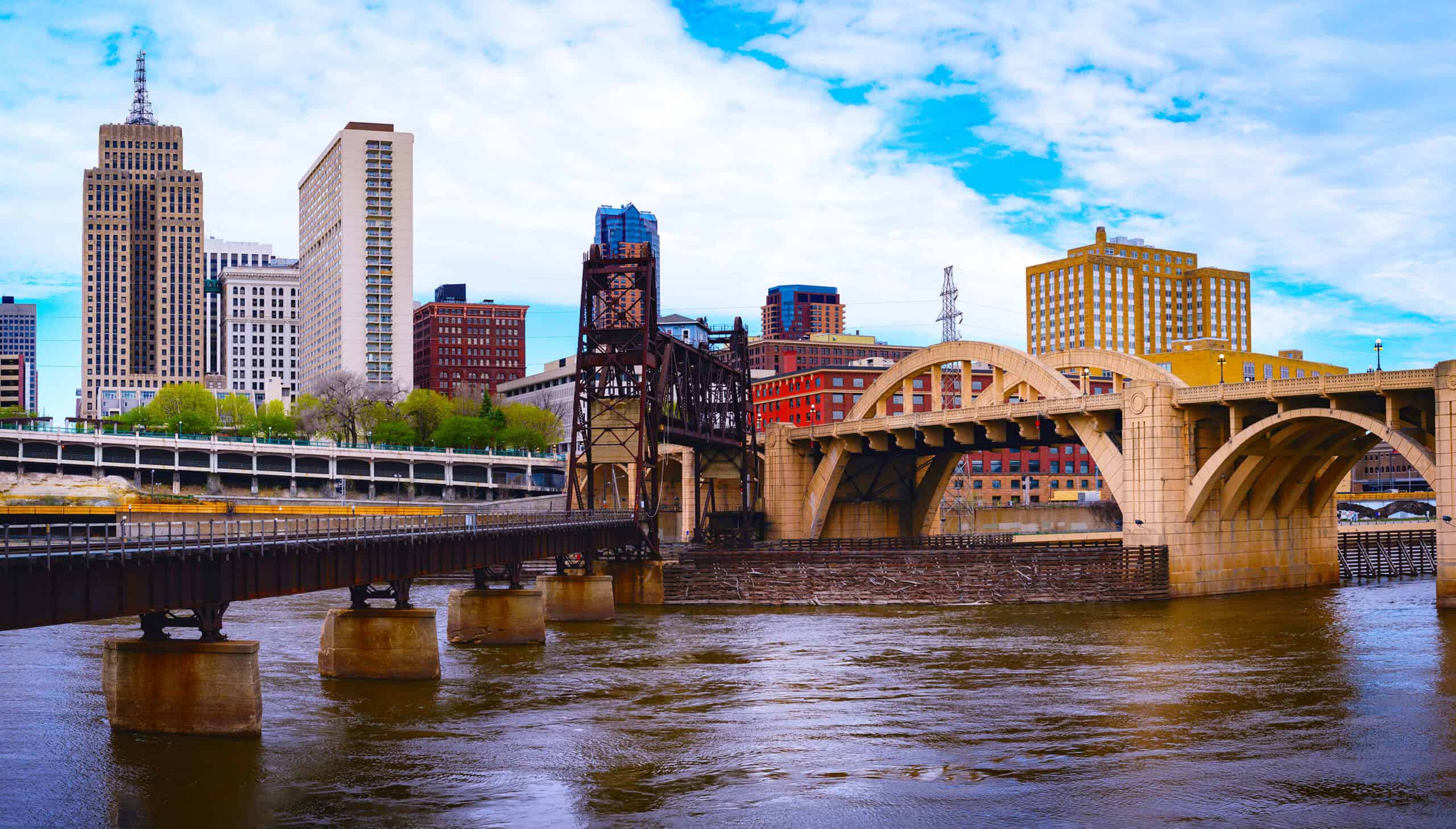 St. Paul, Minnesota skyline with modern and historic buildings, a steel railroad bridge, and a stone arch bridge crossing a wide river.