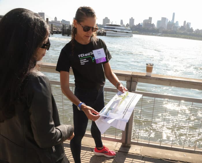 Two women stand by a waterfront railing; one holds and examines a map or plan while the other listens. A ferry and city skyline are visible in the background.