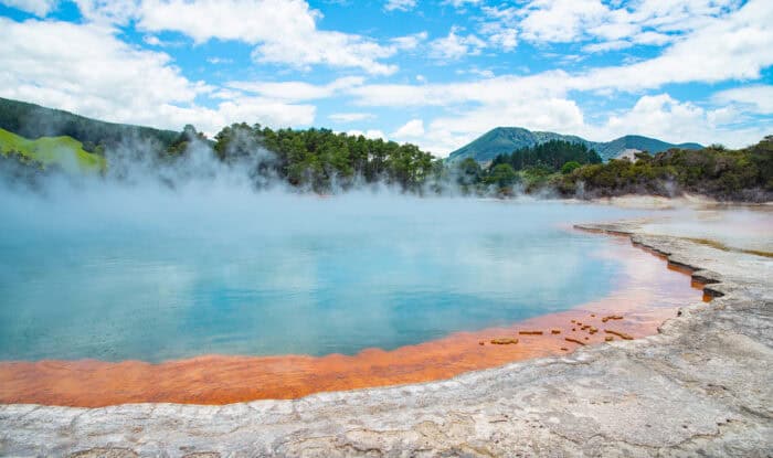 Geothermal hot spring in Rotorua, New Zealand surrounded by rocky edges and green hills under a blue sky.