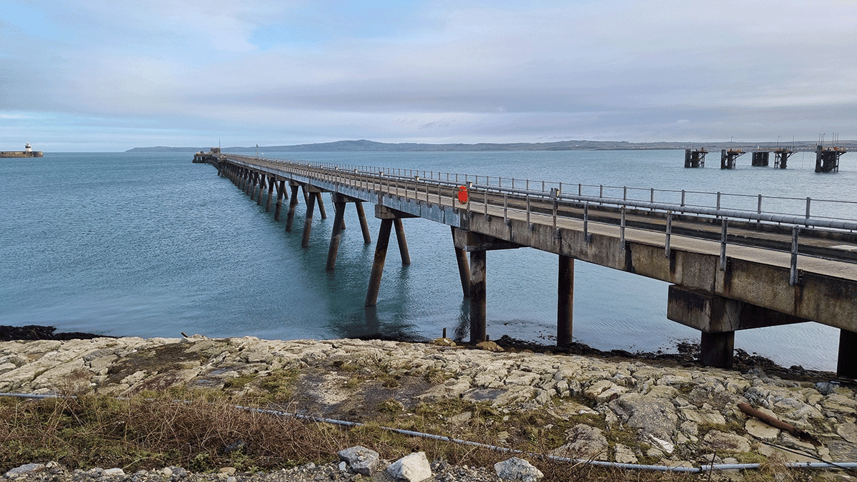 A long concrete pier, part of the Holyhead Jetty inspection by Royal HaskoningDHV, extends over calm blue water, with a rocky shore in the foreground and distant land visible on the horizon under a cloudy sky.