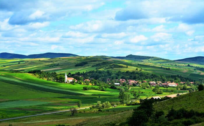 A rural village with red-roofed houses and a white church is surrounded by green rolling hills under a partly cloudy sky.