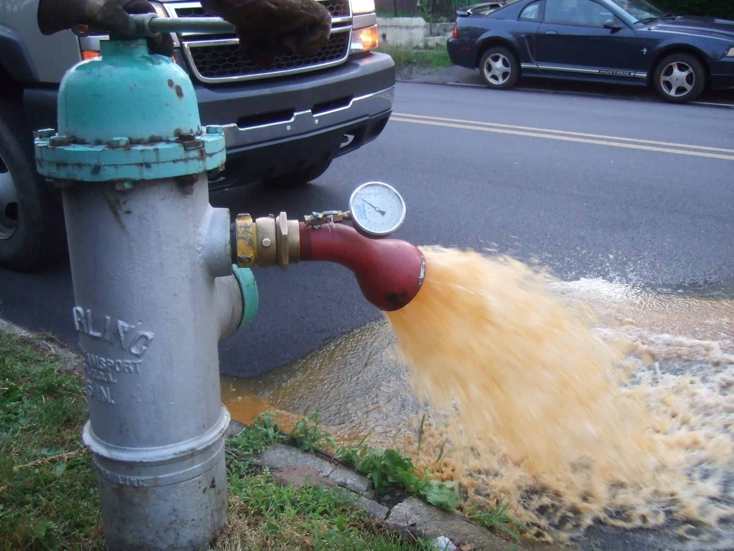 Water flows forcefully from a fire hydrant onto the street during a flushing operation, with a gauge visible on the red attachment. A parked car and part of a truck are seen in the background, all under precise target velocity control.