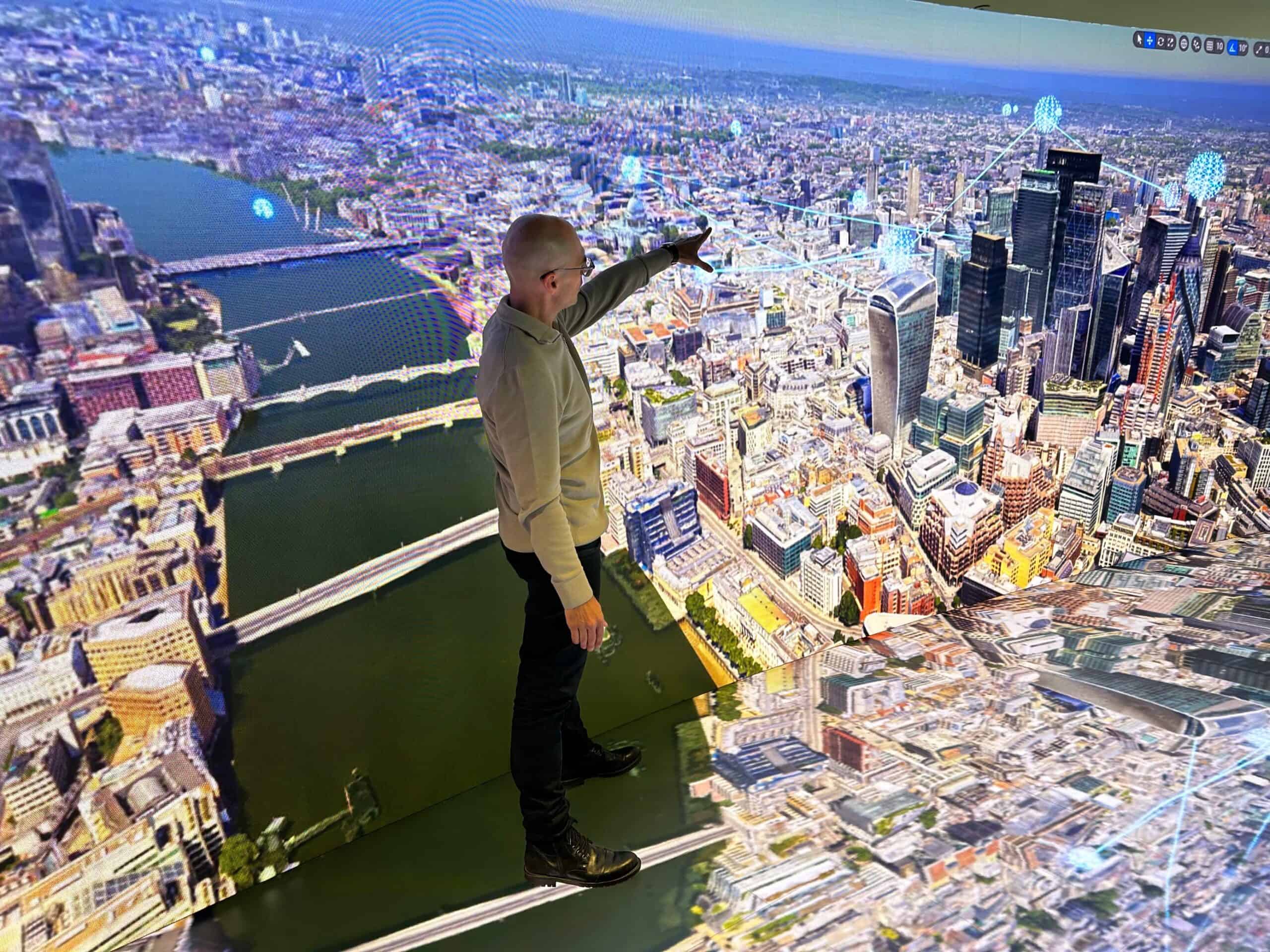 A person gestures towards a large, immersive 3D city map display, depicting a detailed aerial view of an urban landscape with tall buildings, bridges, and a river.