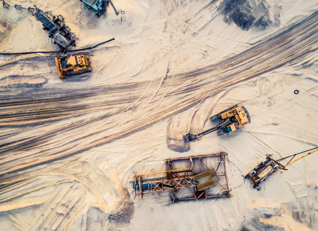 Aerial view of a sandy construction site with heavy machinery and equipment scattered, including excavators and a metal frame structure. Tire tracks are visible on the sand, showcasing detailed area site planning for future development.