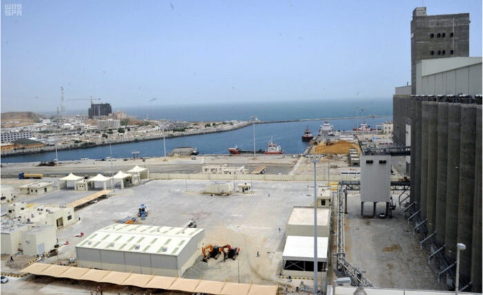 Aerial view of an industrial port area with storage silos, docked ships, and buildings near a body of water under a clear sky.