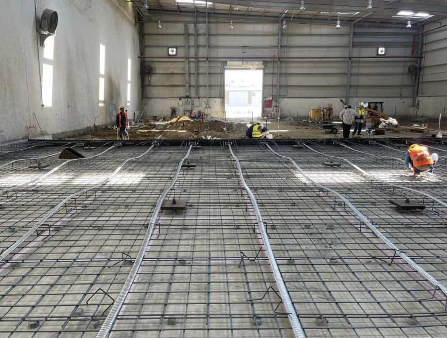 Workers are installing rebar and conduits on a concrete floor inside a large industrial warehouse under construction.