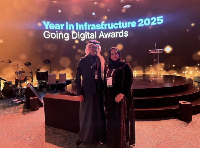 Two people stand in front of a stage with a screen that reads "Year in Infrastructure 2025 Going Digital Awards." Stage lights and musical equipment are visible in the background.