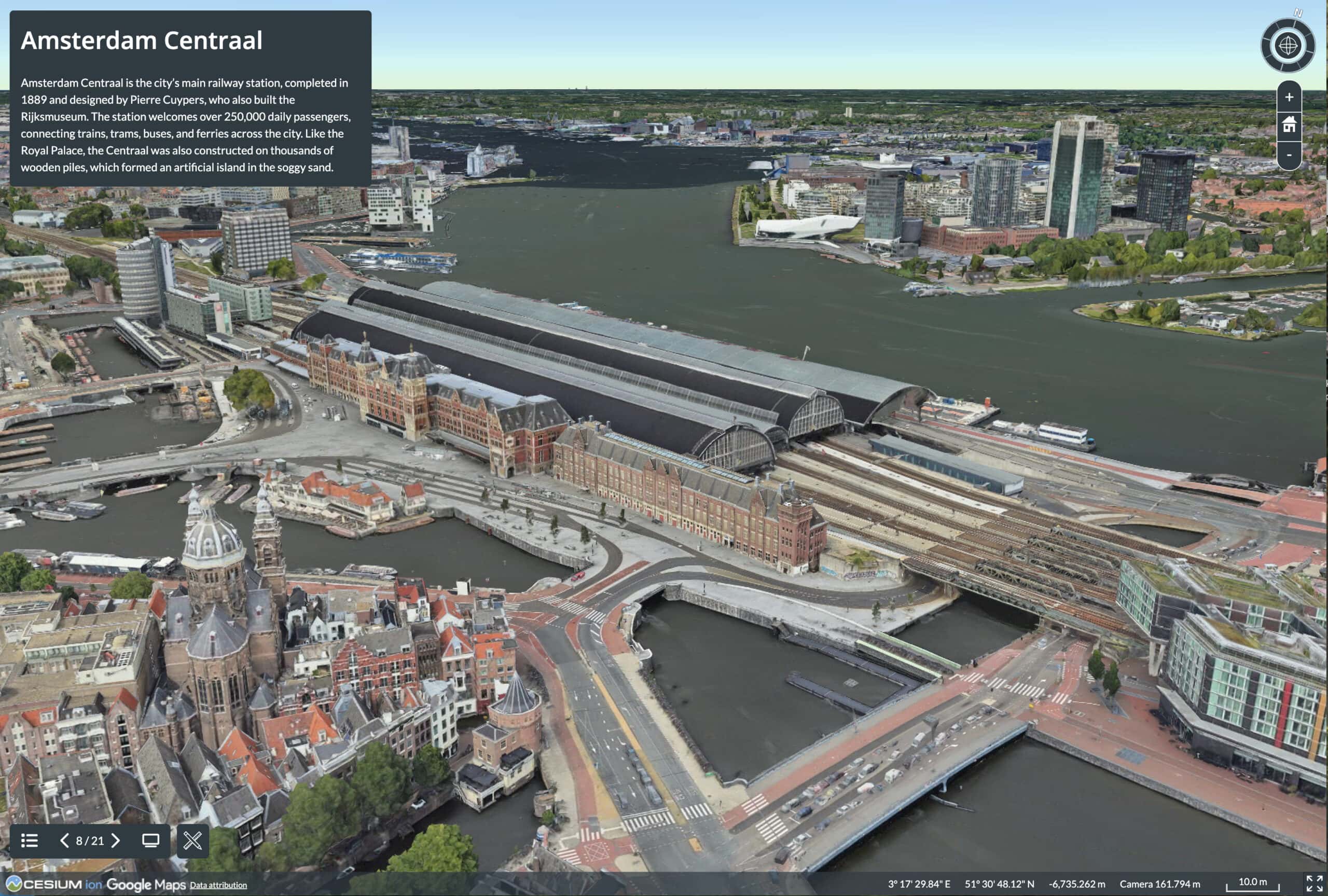 Aerial 3D view of Amsterdam Centraal railway station in the Netherlands, surrounded by water, roads, nearby buildings, and modern high-rises in the background.