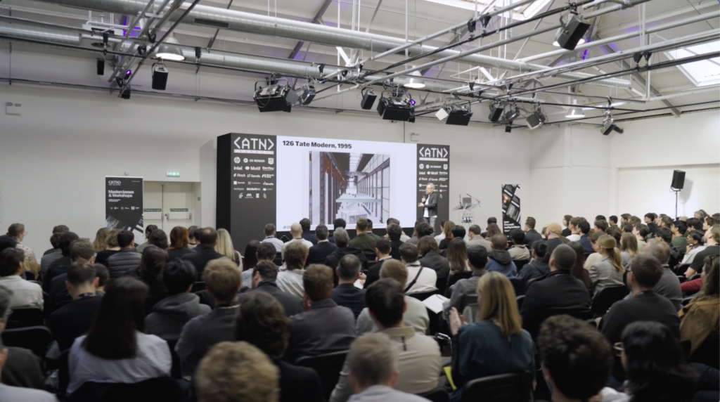 A large audience sits facing a presenter, likely an architect, who stands next to a screen displaying an architectural image with the title "Tate Modern, 1995" in a bright conference room.