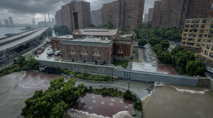 A flooded urban area with rising water surrounding a brick building and nearby basketball courts, highlighting resilient architecture against the backdrop of tall apartment buildings under a cloudy sky.