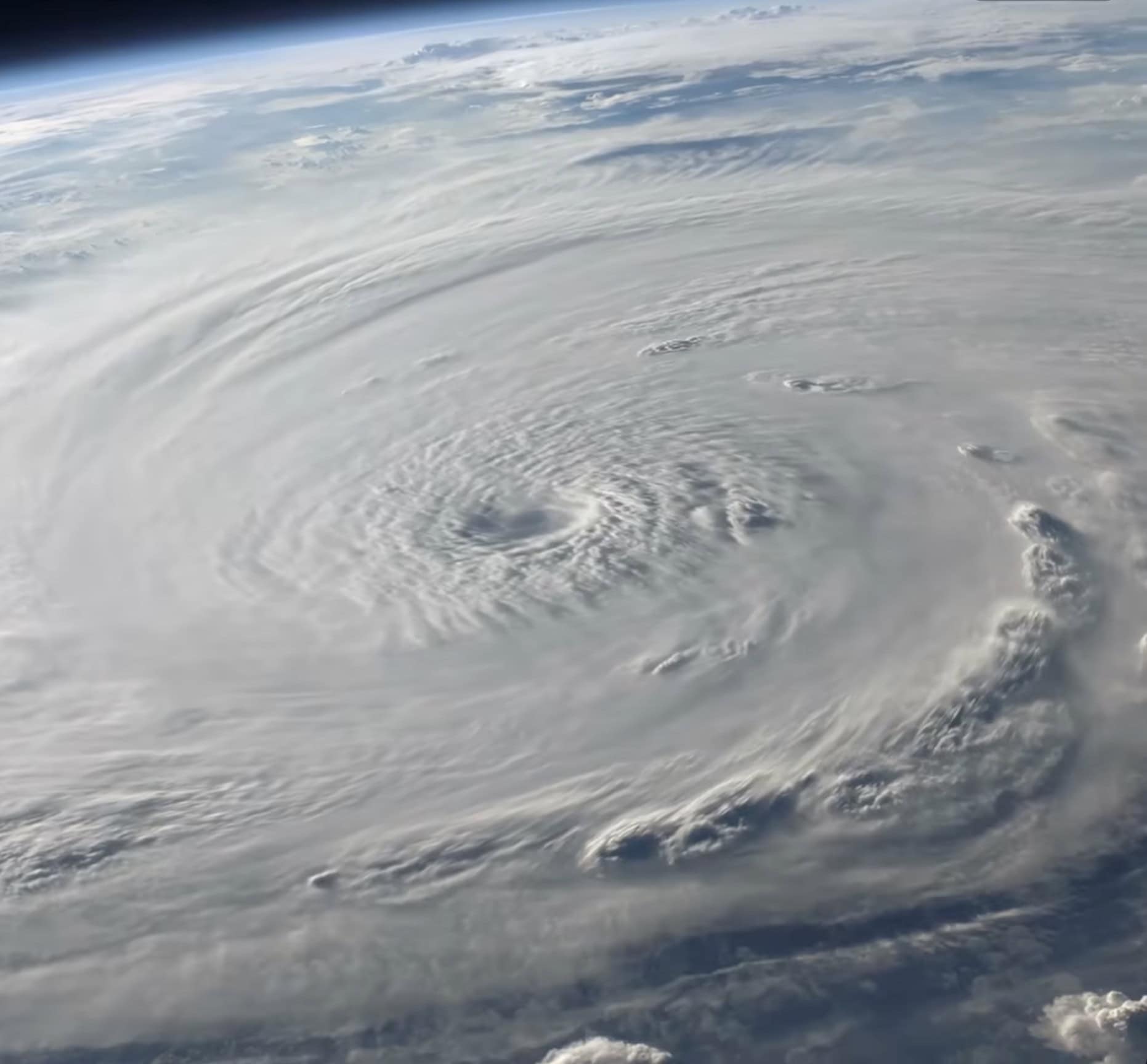 A large hurricane is seen from space, with a visible eye at the center and spiraling clouds extending outward across the Earth's resilient coastline, including parts of New York.