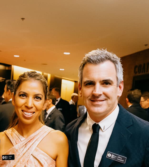 Two people wearing formal attire and name badges smile at an indoor event, with other attendees visible in the background.