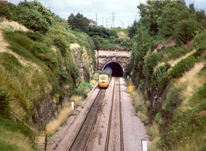 A yellow passenger train exits a tunnel through a grassy hillside, traveling on parallel railway tracks under a partly cloudy sky.