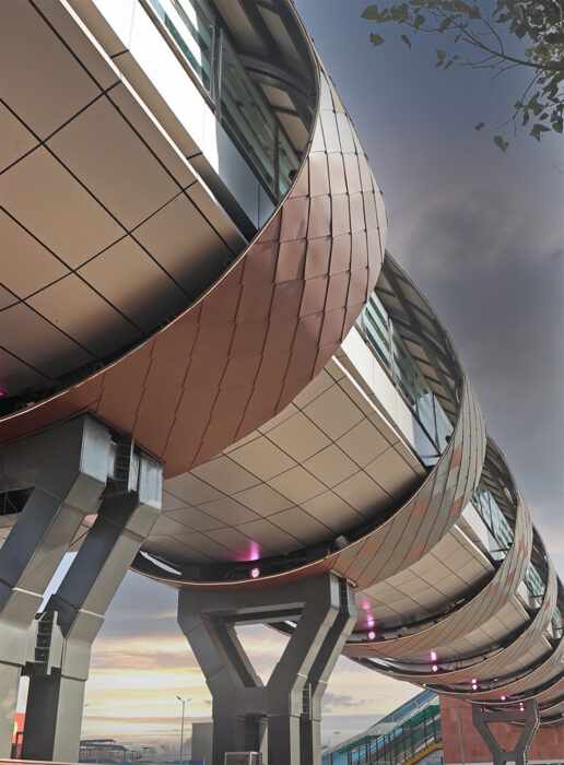 A modern elevated structure by AVS ENGINEERS features curved metallic panels and glass windows, supported by angled steel columns against a cloudy sunset sky—evoking the elegance of Fairmont Palace Udaipur.