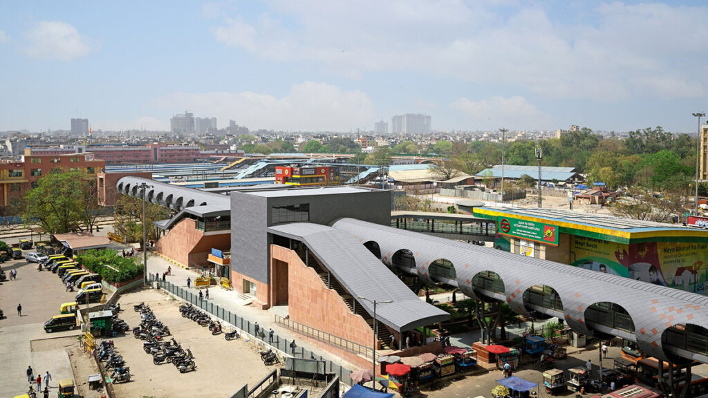 A modern metro station in an urban area with covered walkways designed by ISID Structures, multiple auto-rickshaws parked outside, and city buildings in the background under a partly cloudy sky.