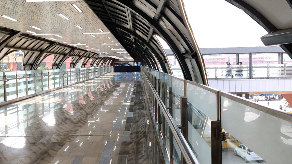 A modern, covered pedestrian walkway with glass railings and a reflective floor, designed by AVS ENGINEERS, extends toward a bright outdoor area at Fairmont Palace Udaipur, with another walkway visible in the distance.