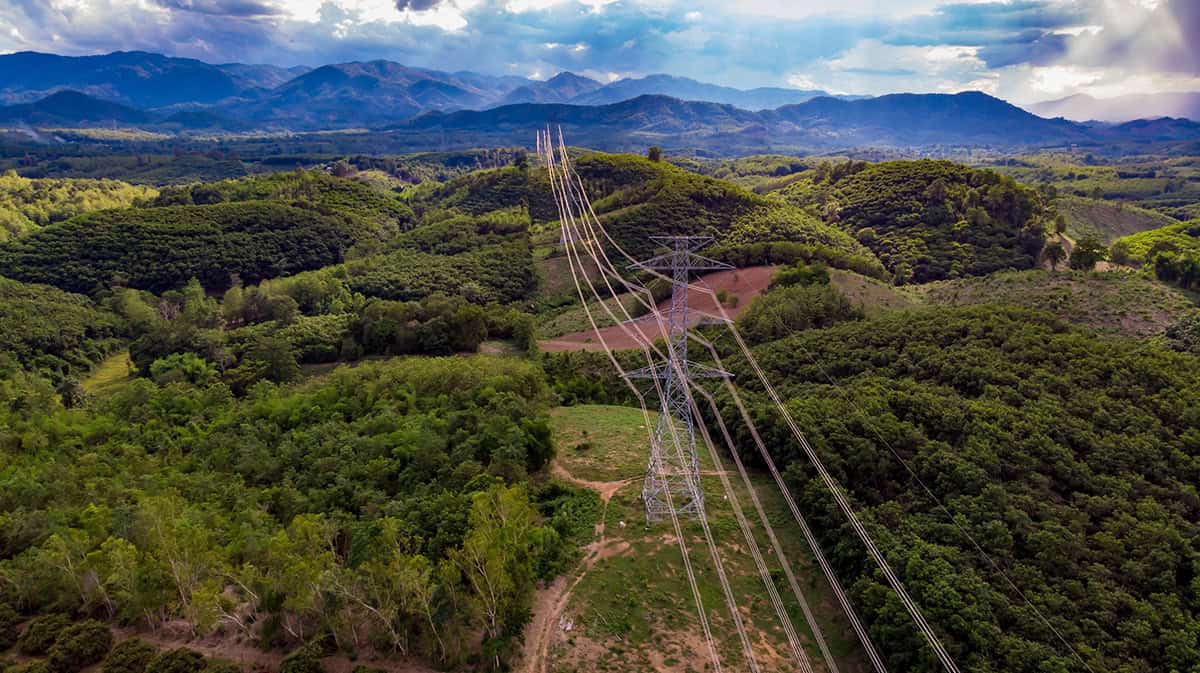 High-voltage transmission tower on a green hillside with several power lines stretching over a valley and distant mountains in view.