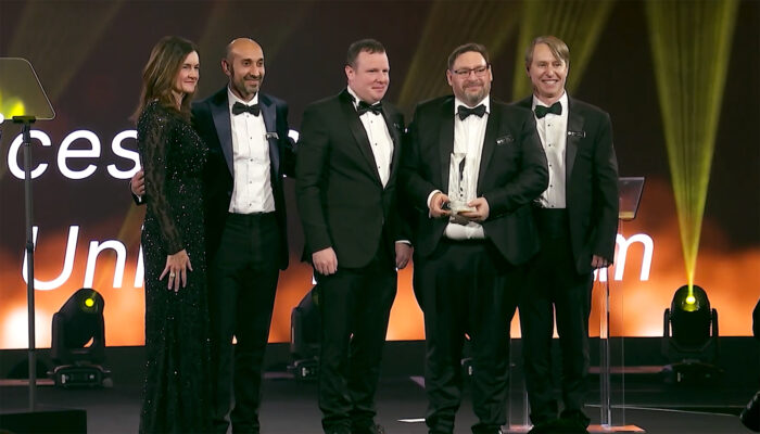 Five people in formal attire stand together at an event; one holds an award. The background is illuminated with warm, golden lights.