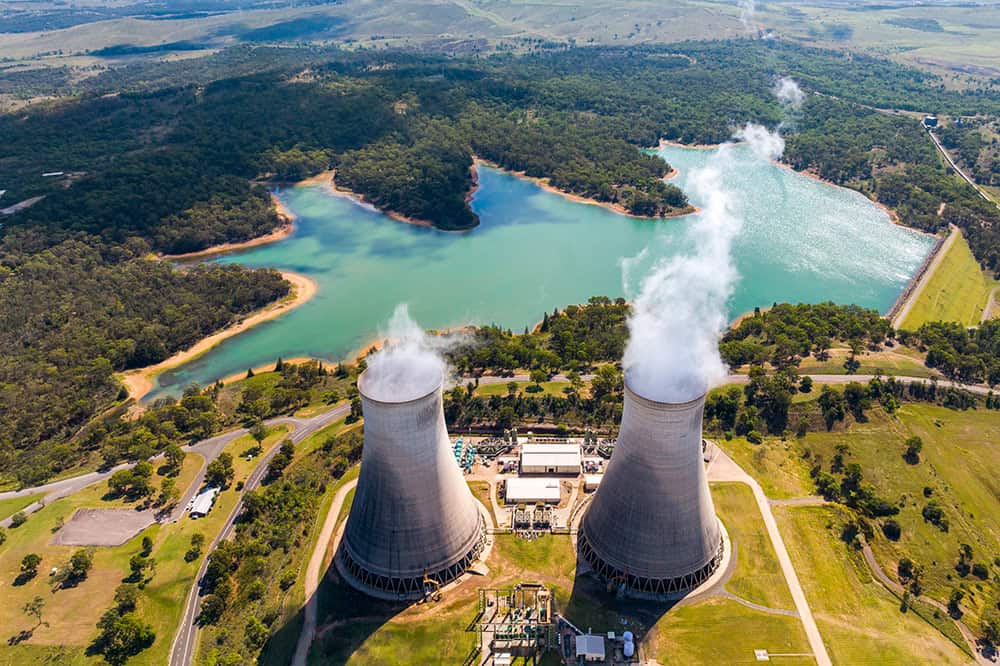 Aerial view of two cooling towers emitting steam at a power plant near a lake, surrounded by trees and grassy landscape.