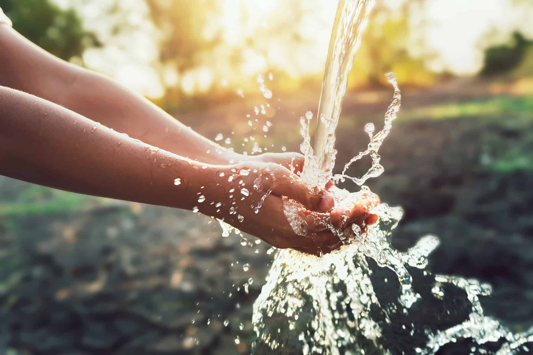 A person holds their hands under a stream of water, with water splashing and sunlight shining in the background—a reminder of the vital role compliance plays in ensuring clean, safe water through efficient water utilities.