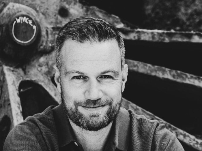 Black-and-white portrait of a bearded man in a collared shirt, smiling at the camera with industrial machinery in the background.