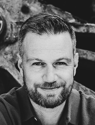 Black and white close-up portrait of a bearded man with short hair, smiling slightly, wearing a collared shirt, with a blurred industrial background.