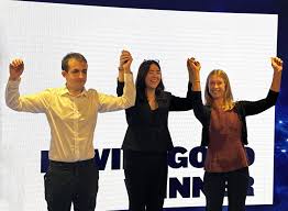 Three people stand in front of a bright sign, holding hands and raising them in celebration, appearing to acknowledge an achievement or award.
